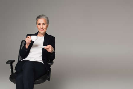 Mature businesswoman holding cup of coffee on office chair isolated on grayの写真素材
