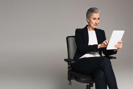 Gray haired businesswoman using digital tablet on office chair isolated on grayの写真素材