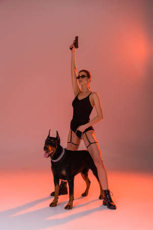 woman in bodysuit holding gun in raised hand near doberman on pink background with yellow lightの写真素材