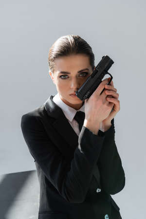 brunette woman holding handgun near face while looking at camera isolated on grayの写真素材