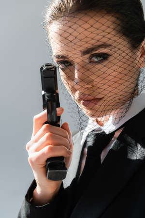 sexy woman in net veil looking at camera while holding handgun isolated on grayの写真素材