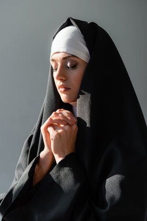 young nun praying with closed eyes isolated on gray