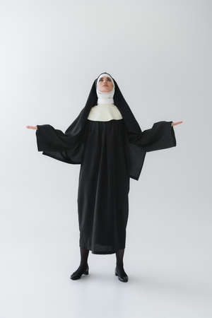 full length view of young nun looking up while praying with open arms on gray backgroundの写真素材