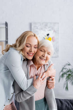Happy woman hugging gray haired mother with closed eyes in living roomの写真素材