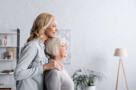 Side view of cheerful woman standing near senior parent in living roomの写真素材