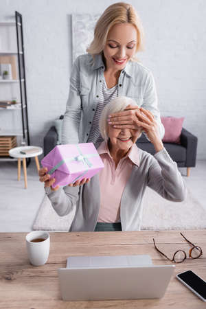 Smiling woman covering eyes to mother while holding present near laptop and tea on tableの写真素材