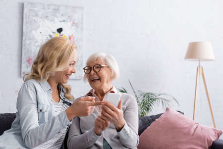 Smiling woman holding mobile phone near daughter on couchの写真素材
