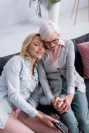Smiling woman holding hand of elderly mother and remote controller on couchの写真素材