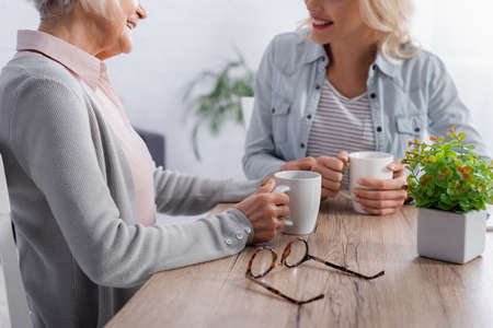 Cropped view of eyeglasses near senior woman holding cup while talking with daughterの写真素材