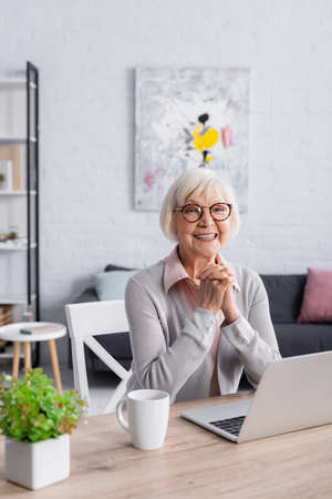 Positive gray haired woman sitting near laptop and cup at home