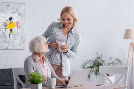 Smiling woman holding cup near mother and gadgets on tableの写真素材