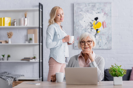 Cheerful elderly woman holding cup near gadgets and daughter on blurred backgroundの写真素材
