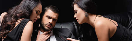 young man looking at camera while sensual women seducing him isolated on black, bannerの写真素材