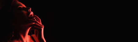 red lighting on seductive woman touching lips isolated on black, bannerの写真素材