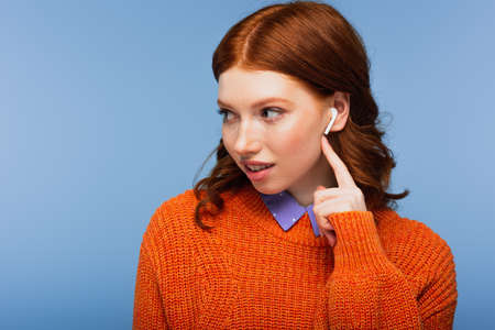 redhead woman listening music while pointing at wireless earphones isolated on blue