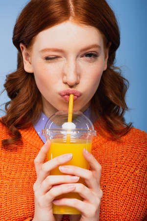 redhead woman in sweater drinking fresh orange juice isolated on blue