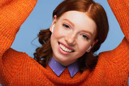 cheerful redhead woman listening music in wireless earphones isolated on blue
