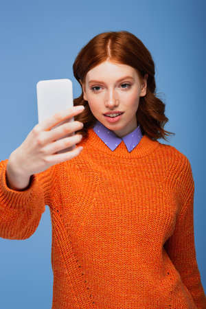 redhead woman in orange sweater taking selfie isolated on blueの写真素材