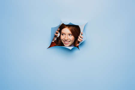 happy redhead young woman looking away on blue ripped backgroundの写真素材