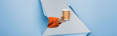 cropped view of young woman holding paper cup with coffee to go on blue ripped paper background, bannerの写真素材
