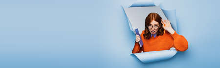 surprised young woman in orange sweater and glasses holding notebook and pencil on blue ripped background, bannerの写真素材