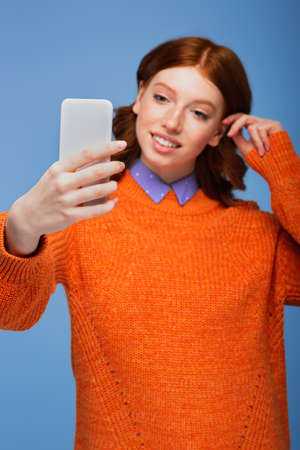 blurred redhead young woman in orange sweater taking selfie on smartphone isolated on blueの写真素材