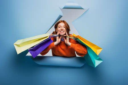 happy redhead woman holding colorful shopping bags on blue ripped paper backgroundの写真素材