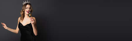 happy young woman in black slip dress and tiara holding glass of champagne on gray, bannerの写真素材