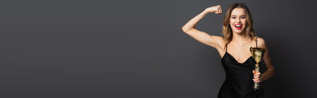 happy young woman in black slip dress holding golden trophy and showing muscle on gray, bannerの写真素材