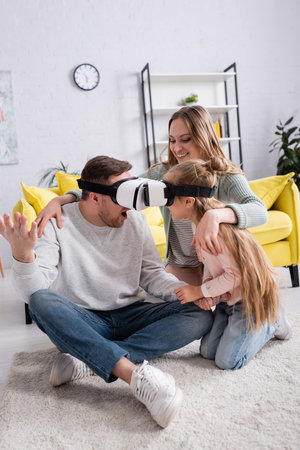 Father and daughter gaming in vr headsets near positive mother on carpet