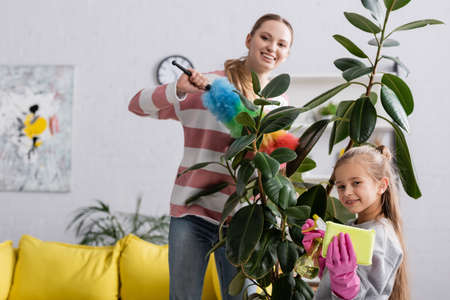 Smiling girl holding detergent and rag near mother and plant