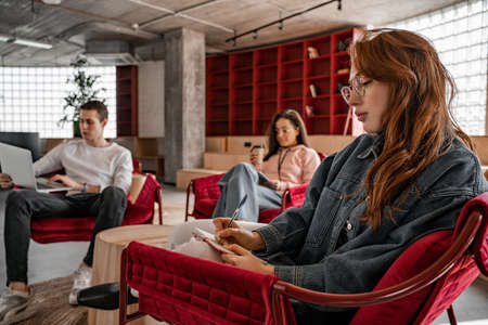 redhead student in glasses writing in notebook near classmates on blurred background, senior 2021