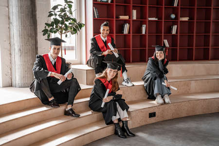 happy interracial students in gowns and caps holding diploma and sitting on stairs, graduation class 2021の写真素材