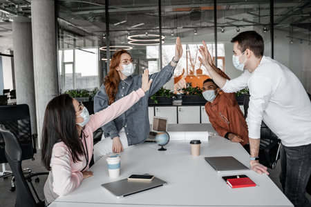 interracial students in medical masks giving high five near gadgets on desk