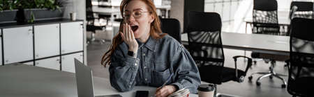 tired redhead student yawning near gadgets and paper cup on desk, bannerの写真素材