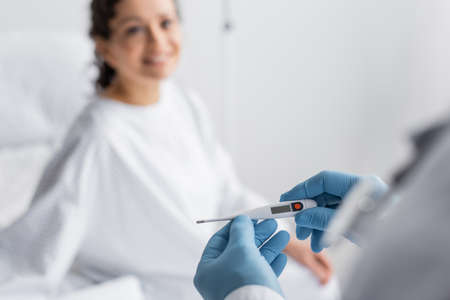 doctor in latex gloves holding thermometer near african american woman smiling on blurred background
