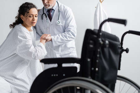 doctor supporting african american woman near wheelchair on blurred foregroundの写真素材