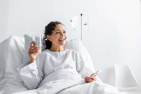happy african american woman smiling while holding glass of water and mobile phone in clinicの写真素材