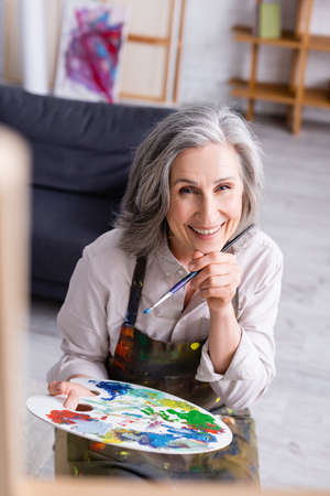 joyful middle aged woman in apron holding paintbrush and palette with colorful paintsの写真素材