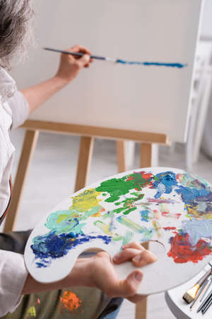 cropped view of middle aged woman holding palette and paintbrush while painting on canvasの写真素材