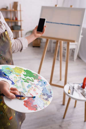 cropped view of mature woman holding palette, paintbrush and smartphone with blank screen near...