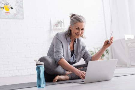 happy middle aged woman sitting in lotus pose on yoga mat and watching online tutorial on laptop and...