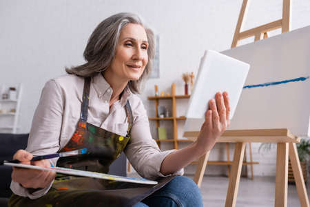 mature woman holding palette and digital tablet while watching tutorial near canvasの写真素材
