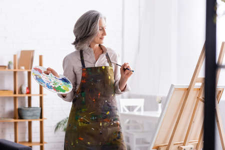 cheerful mature woman in apron holding paintbrush and palette near easel at homeの写真素材