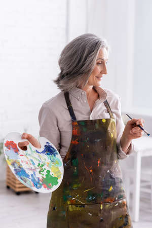 cheerful mature woman in apron holding paintbrush and paletteの写真素材