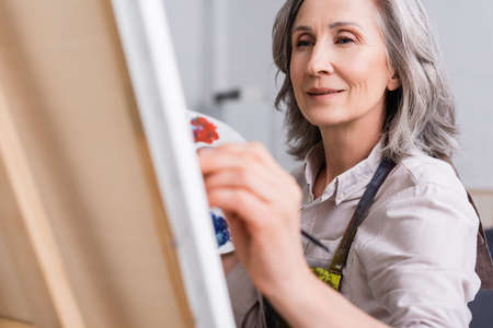 mature woman holding paintbrush while painting on canvas with blurred foregroundの写真素材