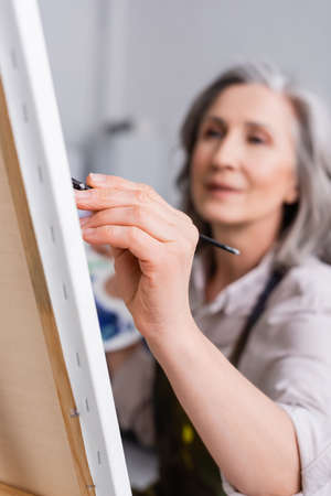 blurred mature woman holding paintbrush while painting with blurred foregroundの写真素材