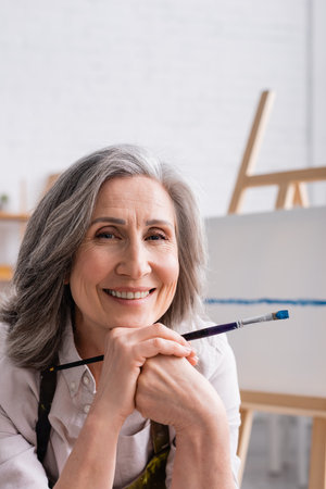 joyful middle aged painter with gray hair holding paintbrushの写真素材