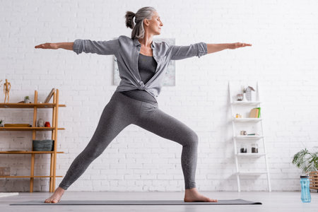 middle aged woman in sportwear standing in warrior pose while practicing yoga at homeの写真素材