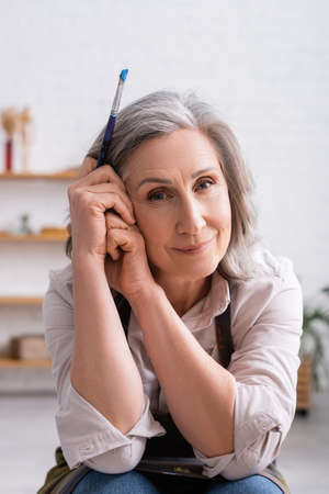 happy middle aged painter with gray hair holding paintbrushの写真素材